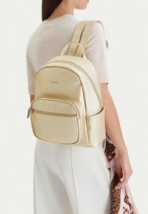 Mujer con top de manga corta beige y pantalones blancos, llevando una mochila metálica dorada con bolsillo frontal con cremallera y compartimento lateral.