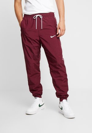Osoba ubrana w bordo spodnie dresowe Nike z białym sznurkiem i czerwonymi bocznymi paskami, do tego białe buty sportowe Nike z zielonym znakiem Swoosh.
