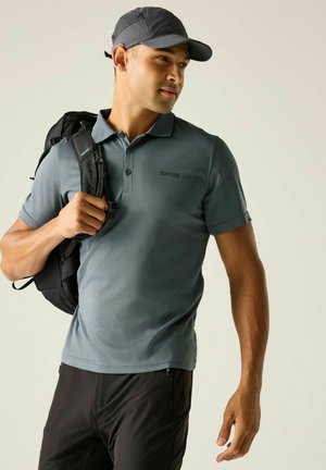 Ein Mann trägt ein graues Poloshirt und eine schwarze Kappe, trägt einen schwarzen Rucksack über einer Schulter und schaut leicht zur Seite.