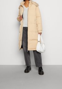 Manteau en duvet beige avec capuche, poignets côtelés et fermeture à pression. Porté avec un pull gris clair, un pantalon large gris et des bottes noires.