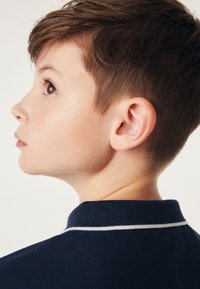 Tumši zilas polo krekls ar ribinātu apkakli un balto apdari, ko valkā bērns sānu skatījumā, izceļot īsus matus un ausi.