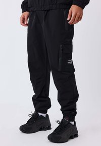 Persona che indossa pantaloni cargo neri con una tasca laterale con zip e scarpe Nike Shox nere, in piedi su uno sfondo bianco.