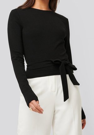 Top negro de manga larga hecho de tela suave, con detalle de cintura atada, escote redondo y mangas ajustadas. Llevado con pantalones blancos.