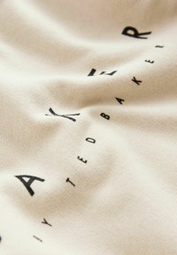 Tejido de color beige claro con una superficie texturizada que presenta texto impreso en negro que dice "BAKER". El diseño es simple y moderno.