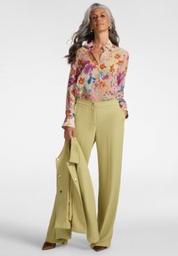 Kvinde med langt gråt hår iført en flerfarvet blomstret bluse og lysegrønne bukser, der holder en matchende lysegrøn blazer.