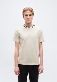 Ανοιχτό μπεζ κοντομάνικο μπλουζάκι με στρογγυλή λαιμόκοψη, διακοσμημένο με μαύρη αντίθετη επένδυση και μικρό λογότυπο στο κέντρο της λαιμόκοψης.