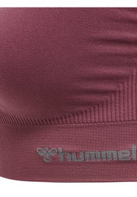 Zbliżenie na teksturowaną bordową tkaninę z prążkowanymi detalami i szarym logo "hummel" na dolnej krawędzi ubrania.