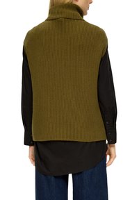 Olijfgroene geribbelde gebreide vest met een hoge kraag, gedragen over een zwart langemouwshirt, met een relaxte pasvorm en een rechte zoom.