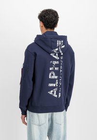 En person med krøllete hår iført en navy blå hettegenser med kamuflasje "Alpha Industries"-tekst på ryggen, kombinert med lyseblå jeans, snur ryggen til.
