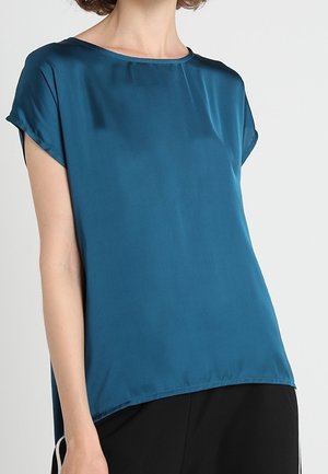 Femme portant un chemisier en satin bleu sarcelle à manches courtes avec un col rond et un pantalon noir sur un fond uni.