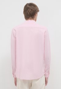 Person von hinten betrachtet, der ein hellrosa Langarmshirt und eine cremefarbene Hose vor einem schlichten weißen Hintergrund trägt.