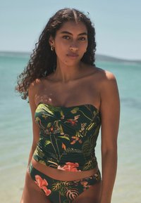 Bikini set s cvjetnim uzorkom koji ima zelenu podlogu, nabran gornji dio i vibrantne cvjetne motive. Glatka tkanina s prilagođenim dizajnom i minimalnim pokrićem.