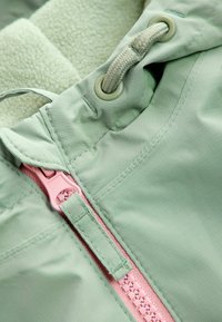 Giacca verde chiaro con morbido rivestimento in pile crema, zip rosa a contrasto e cordino regolabile nel collo.