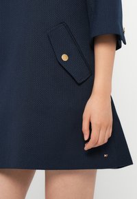 Marinefarget teksturert kjole med en stor klafflomme med en gullknapp og en liten logodetalj på kanten. Korte ermer og utsvingt design.