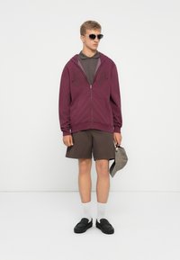 Ung mand iført solbriller, en lilla zip-up hoodie, brune shorts, hvide sokker, sorte sko, og holder en grå kasket mod en ensfarvet hvid baggrund.