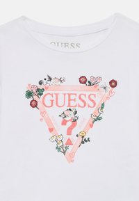 Weißes Baumwoll-T-Shirt mit einem rosa glitzernden "GUESS"-Logo in einem Dreieck, verziert mit bunten Blumenillustrationen und kleinen Tiergrafiken.