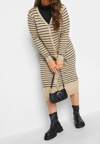 Béžové pruhované šaty na zapínání s výstřihem do V, knoflíky vpředu a žebrovanými manžetami; doplněné černými kotníkovými botami a malou černou kabelkou.