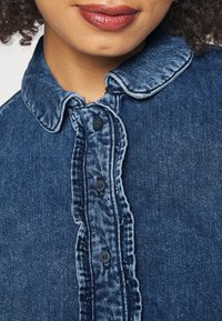 Camicia in denim blu scuro con colletto arrotondato, cinque bottoni lungo il davanti e un tessuto leggermente testurizzato con dettagli di cuciture sfilacciate.
