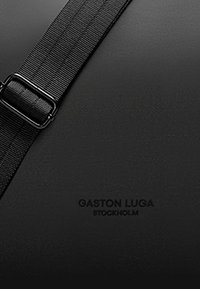 Zaino nero con superficie texturizzata, tracolla nera regolabile con fibbia in metallo e nome del brand in rilievo "Gaston Lugo Stockholm."