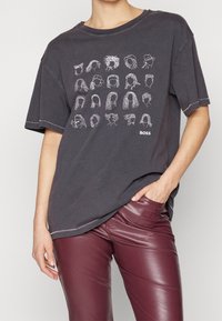 Person klädd i en mörkgrå T-shirt med 24 vita linjetecknade ansikten och texten "BOSS", tillsammans med vinröda läderbyxor.