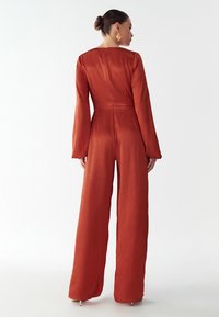 Rudos spalvos kombinezonas su ilgomis rankovėmis, plačiomis kojomis ir prigludusia kūno dalimi. Pagamintas iš tekstūruoto audinio, turi užtrauktuką gale ir minimalistinį dizainą.