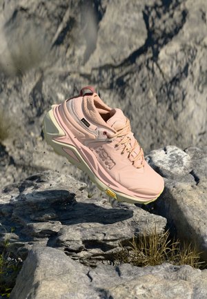 Różowy but do biegania po szlaku z grubą podeszwą, skaczący nad skalistym terenem na zewnątrz.