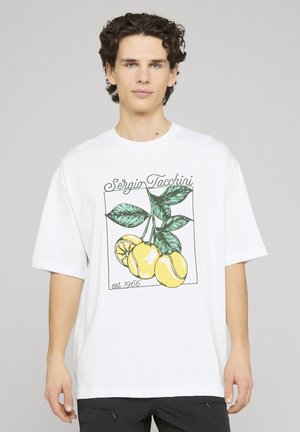 Ung mand iført en hvid t-shirt med en gul citron og en tennisbold grafik samt teksten "Sergio Tacchini est. 1966" foran.