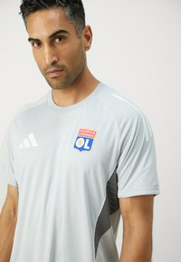 Grå treningstrøye med korte ermer, med logo av Olympique Lyonnais og mønstrede detaljer på sidene og skuldrene.