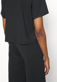 Svart ribbad kortärmad topp med avslappnad passform, med en rundad fåll och en liten logotypdetalj på sidan, kombinerad med matchande mörka byxor.