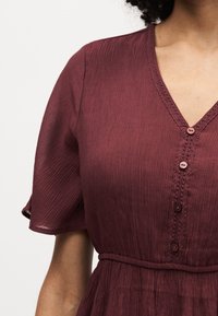 Γυναίκα με μπορντό υφασμάτινο φόρεμα με κοντά μανίκια, V-λαιμόκοψη, κουμπιά μπροστά και εφαρμοστό ζωνάκι στη μέση.