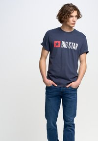 Tumši zils kokvilnas T-krekls ar sarullētām piedurknēm, uz kura ir sarkans kvadrāts un balti uzraksts "BIG STAR". Kombinēts ar ziliem džinsiem.