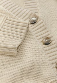Cremfarbener gestrickter Cardigan mit gerippten Bündchen und Kragen, der auf der Vorderseite drei braune Verlaufsknöpfe hat.