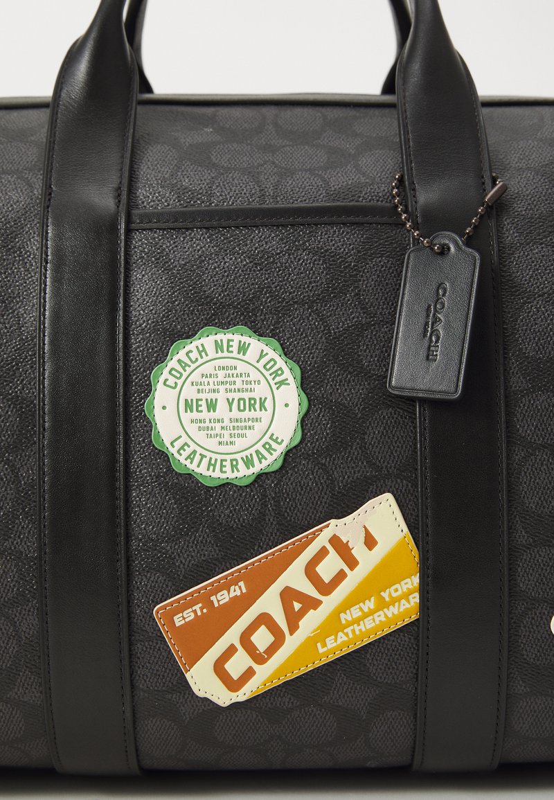 Black leather duffel bag with embossed pattern, featuring green and orange travel stickers and a black tag attached by a chain.