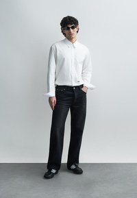 Λευκό πουκάμισο με μακριά μανίκια, σκούρο τζιν, μαύρα αθλητικά παπούτσια με λευκές ρίγες. Το μοντέλο φοράει σκούρα γυαλιά ηλίου. Απλός, μοντέρνος σχεδιασμός.