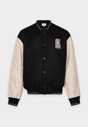 Veste universitaire noire à manches crème, avec des boutons-pression, des bordures côtelées à rayures et un écusson avec la lettre K sur la poitrine.