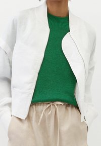Giacca bomber bianca con polsini a costine, indossata sopra un maglione verde texturizzato, abbinata a pantaloni beige chiaro con coulisse.