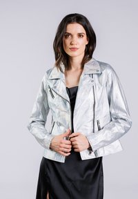 Veste de moto argentée irisée avec une coupe courte, poches zippées et col cranté, superposée à une robe noire.