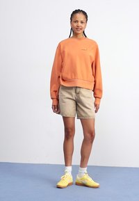 Orange sweatshirt med ribbede manchetter og kant, parret med beige shorts. Modellen bærer gule sneakers og hvide strømper. Baggrunden er ensfarvet.
