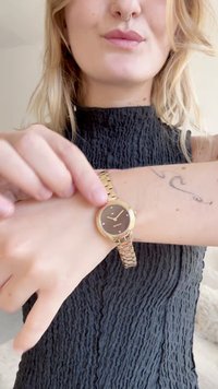 Zlatý náramkové hodinky s kulatým tmavým ciferníkem, třemi malými akcentovými značkami a spojeným kovovým páskem, vystavené na zápěstí.