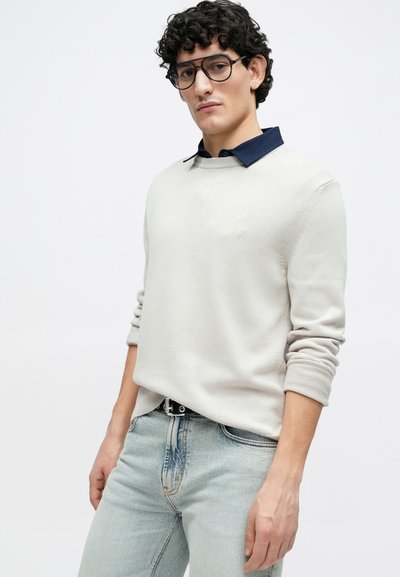 Pull gris clair avec un col bleu marine, doté de poignets côtelés. Associé à un jean en denim bleu clair et des lunettes noires rectangulaires.