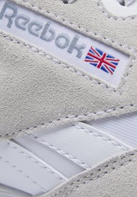 Vitt sportskor med överdel i mocka, läderdetaljer och sydda detaljer. Reebok-logotyp med brittisk flaggetikett synlig.