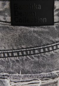 Étiquette en cuir noir avec "Bershka Denim Collection" sur tissu denim gris usé avec coutures visibles et bord effiloché.