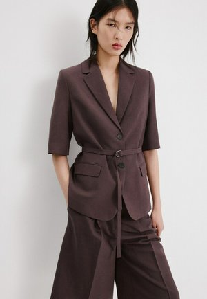Młoda kobieta o prostych czarnych włosach nosi ciemnobrązową marynarkę z paskiem, o długości rękawów do łokcia, oraz dopasowane szerokie spodnie.