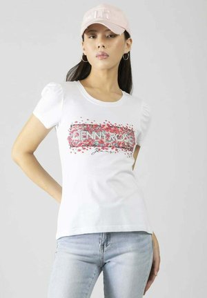 Donna che indossa una T-shirt bianca con grafica floreale, jeans azzurri, cappellino rosa e orecchini a cerchio su sfondo grigio uniforme.