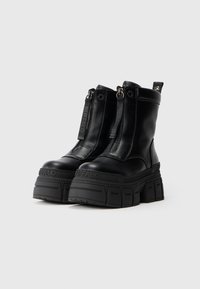 Botas de pele pretas com solas chunky proeminentes. Apresentam zíperes duplos na frente e uma marca gravada na lateral. Textura mate.