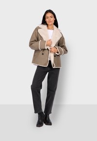 Giacca di shearling marrone con colletto in pelliccia crema, bottoni doppi, e maniche lunghe, abbinata a jeans neri e stivali.