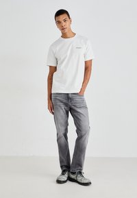 Hvit kortermet t-skjorte med en liten logo, sammen med grå smale jeans og lyse sko, står mot en nøytral bakgrunn.