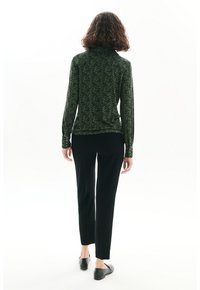 Haut à manches longues vert à motifs avec un col montant, associé à un pantalon noir ajusté et des chaussures noires à enfiler. La texture semble lisse.