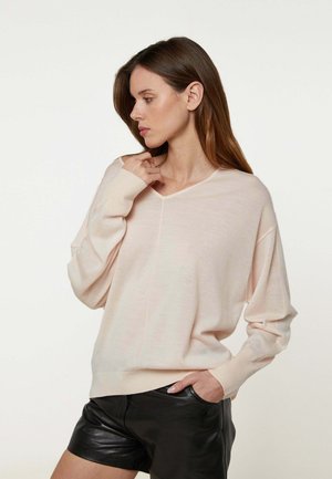 Vrouw met lang bruin haar, gekleed in een licht beige trui met V-hals en zwarte leren korte broek, poserend met één hand in de zak en de andere bij haar nek.