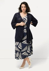 Kimono azul marino de corte abierto con mangas de 3/4, que presenta patrones decorativos. Debajo, un vestido con estampado de hojas en azul marino y crema. Tacones puntiagudos beige.